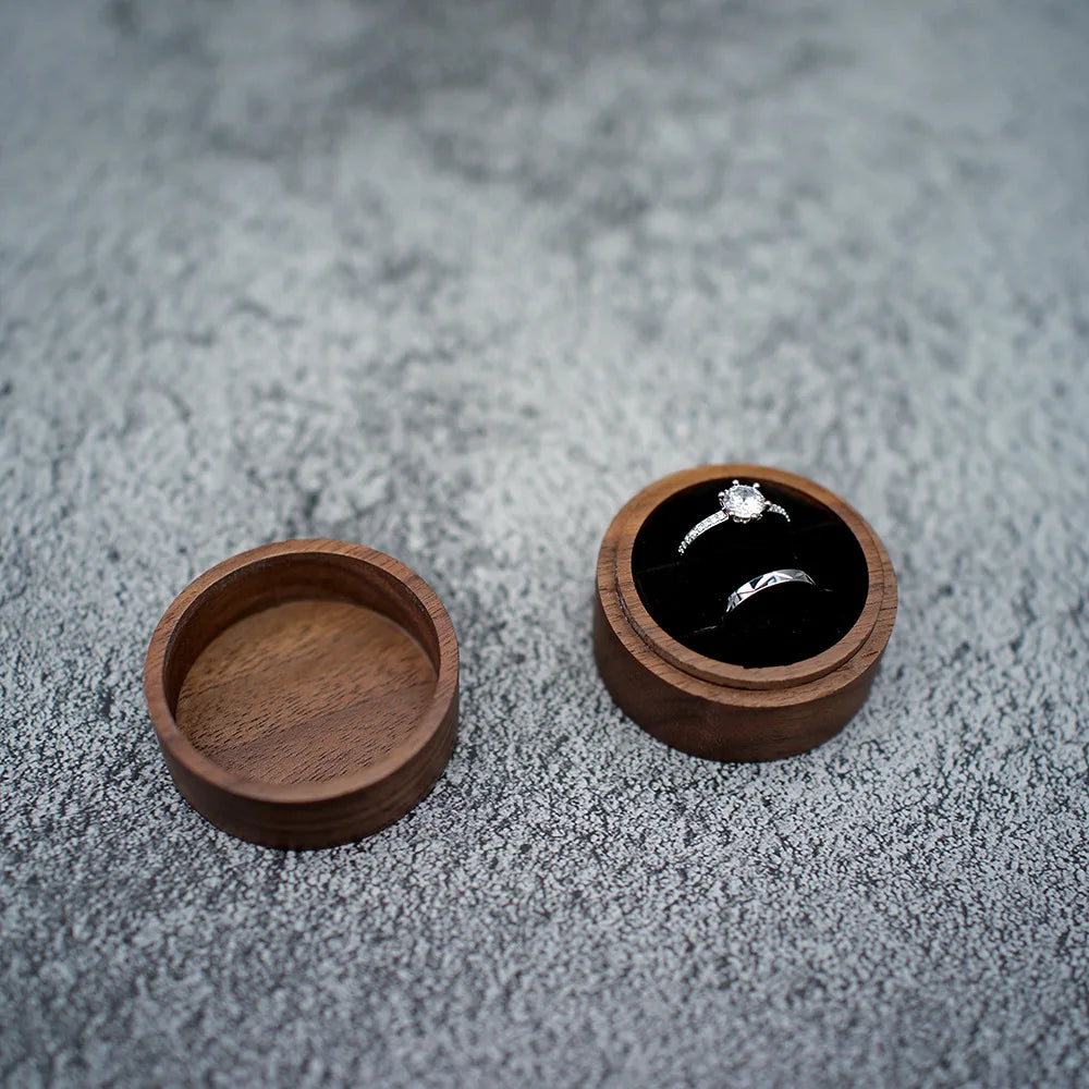 Boîte à bague de fiançailles ronde en bois avec couvercle séparé, ouverte, contenant deux bagues argentées sur velours noir.