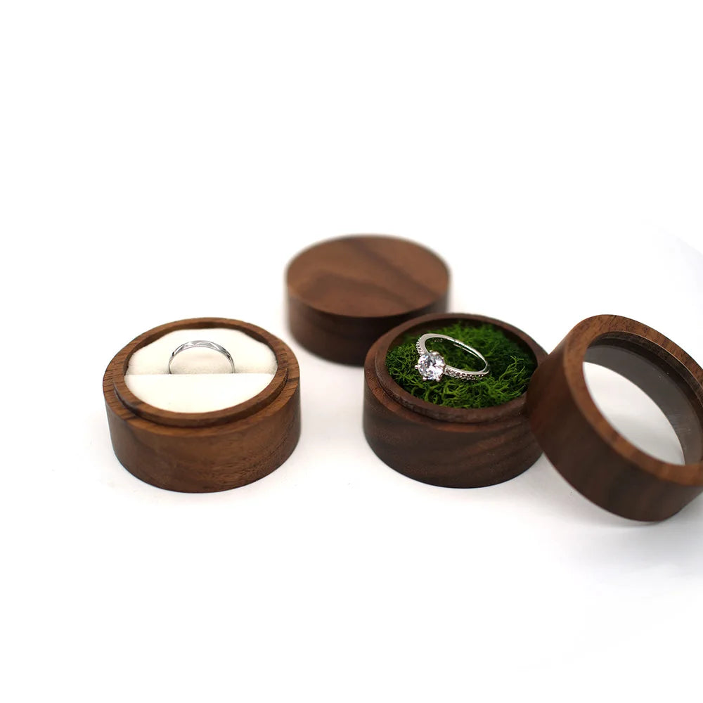 Boîtes à bague rondes en bois, l'une avec un anneau simple sur tissu blanc, l'autre avec un anneau à diamant sur mousse verte