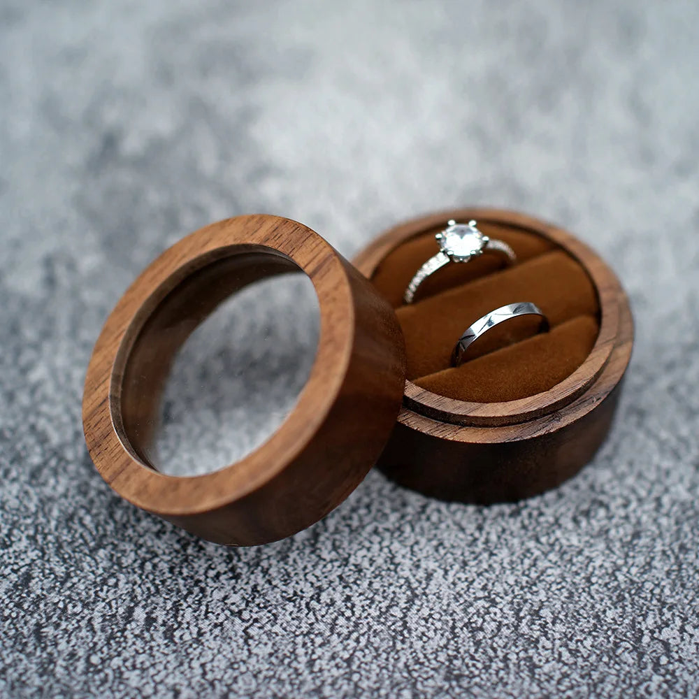 Boîte à bague de fiançailles ronde en bois sombre, ouverte, avec deux bagues sur un intérieur en velours marron.