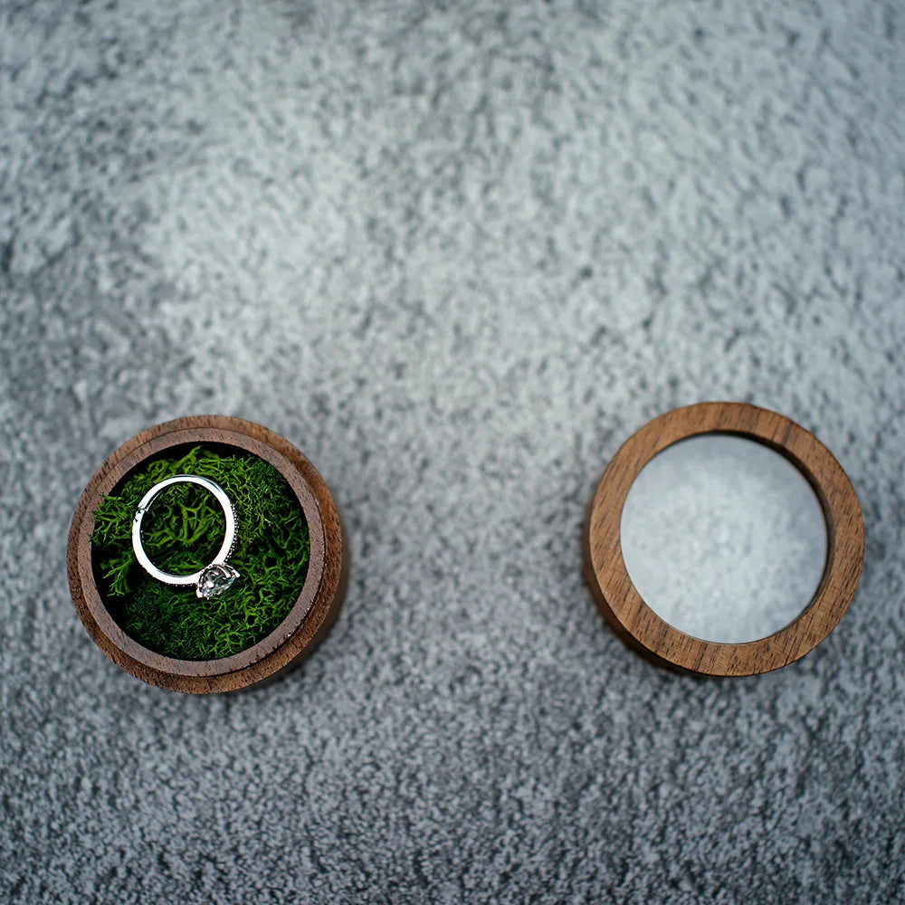 Boîte à bague ronde en bois foncé, ouverte sur un lit de mousse verte avec une bague en argent. Le couvercle est à côté.