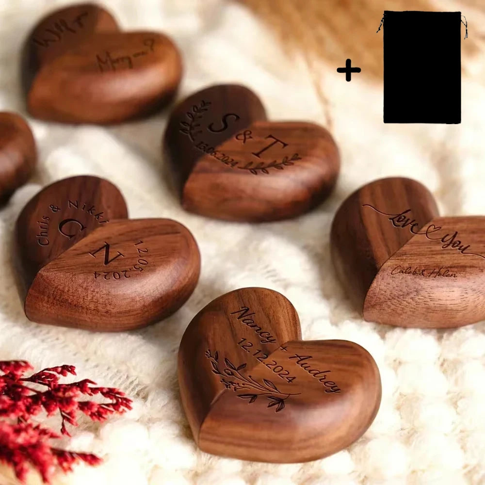 Boîtes à bagues en bois de noyer en forme de cœur, gravées de noms et de dates, posées sur un tissu clair.