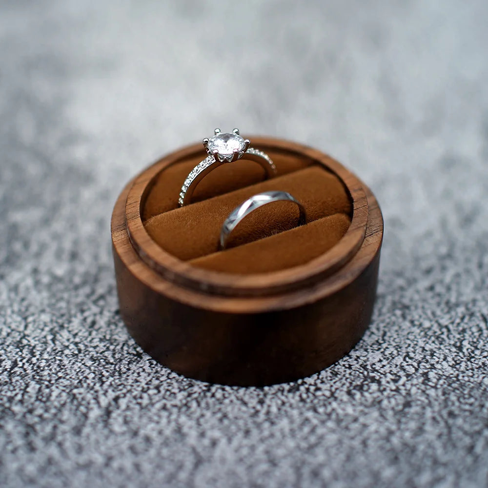 Boîte à bague de fiançailles ronde en bois avec un intérieur en tissu marron, contenant deux anneaux.