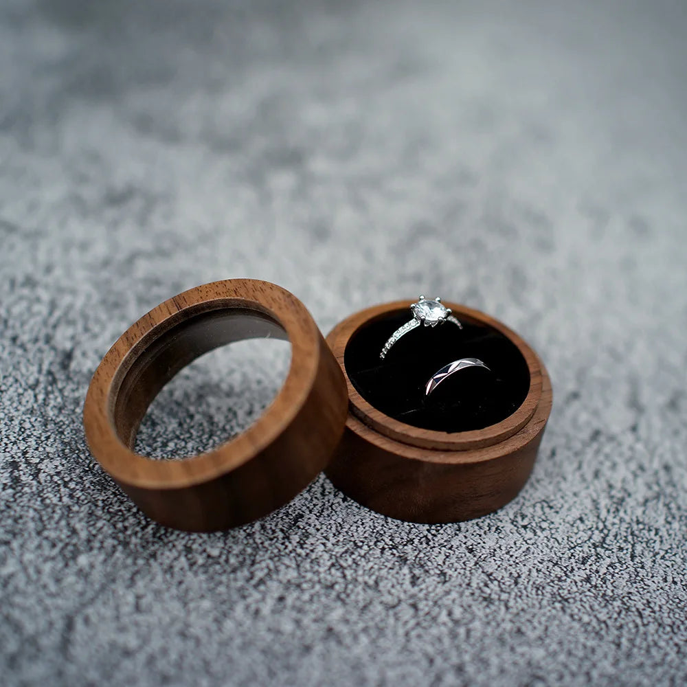 Boîte à bague de fiançailles ronde en bois avec couvercle, intérieur en tissu noir, contenant deux anneaux.