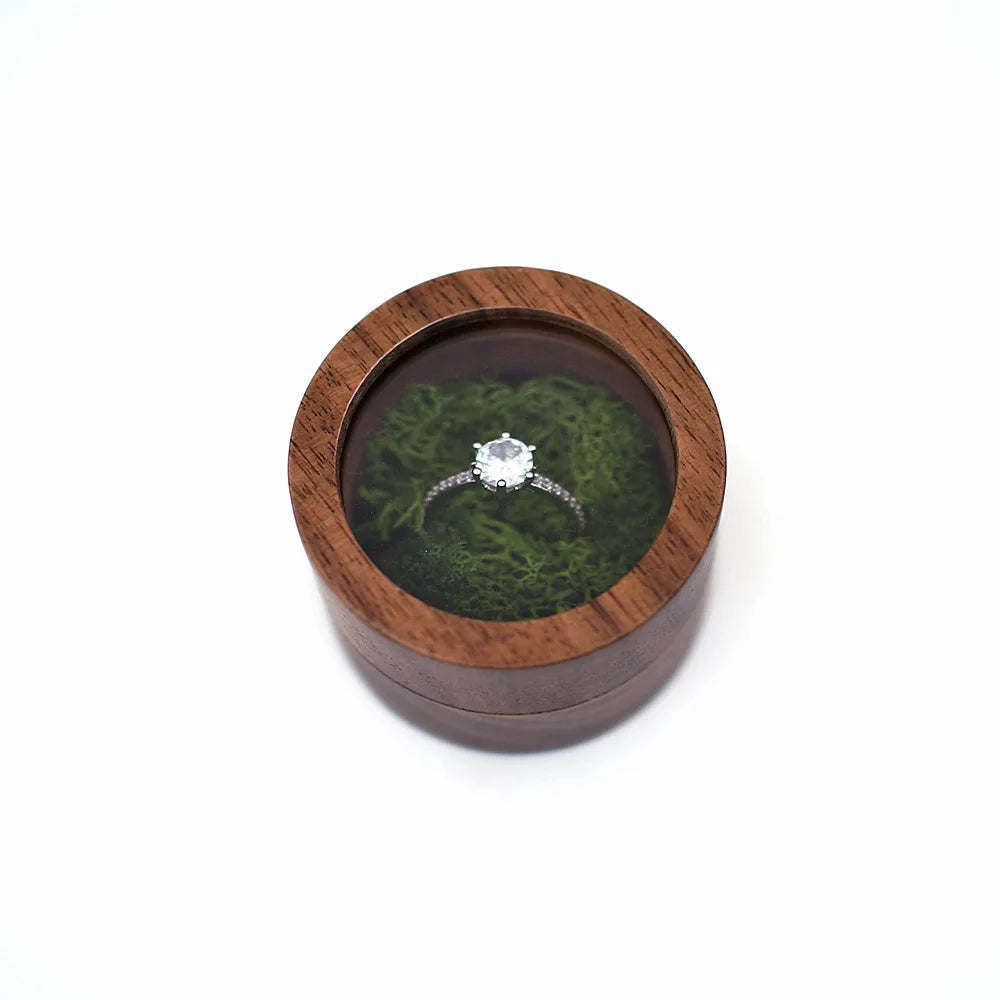 Boîte à bague de fiançailles ronde en bois avec couvercle transparent, contenant une bague argentée sur un lit de mousse vert