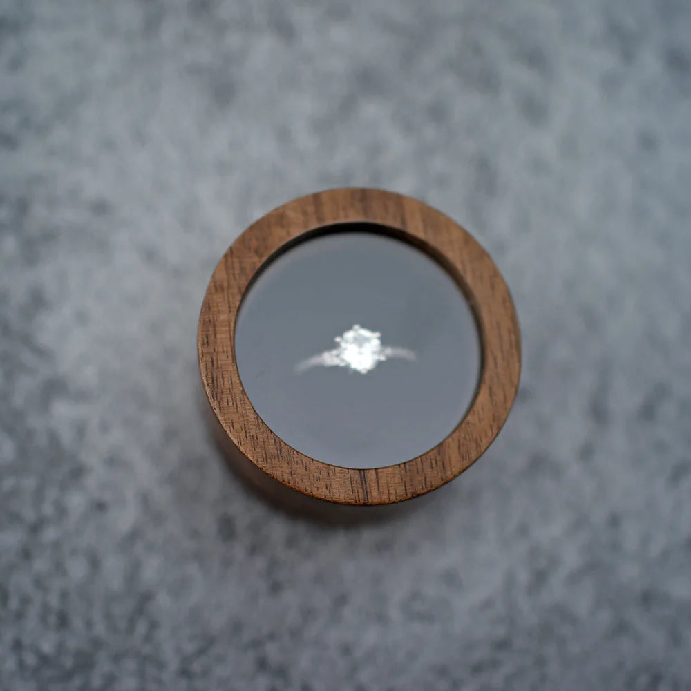 Boîte à bague de fiançailles ronde en bois sombre, avec couvercle transparent révélant une bague en argent.