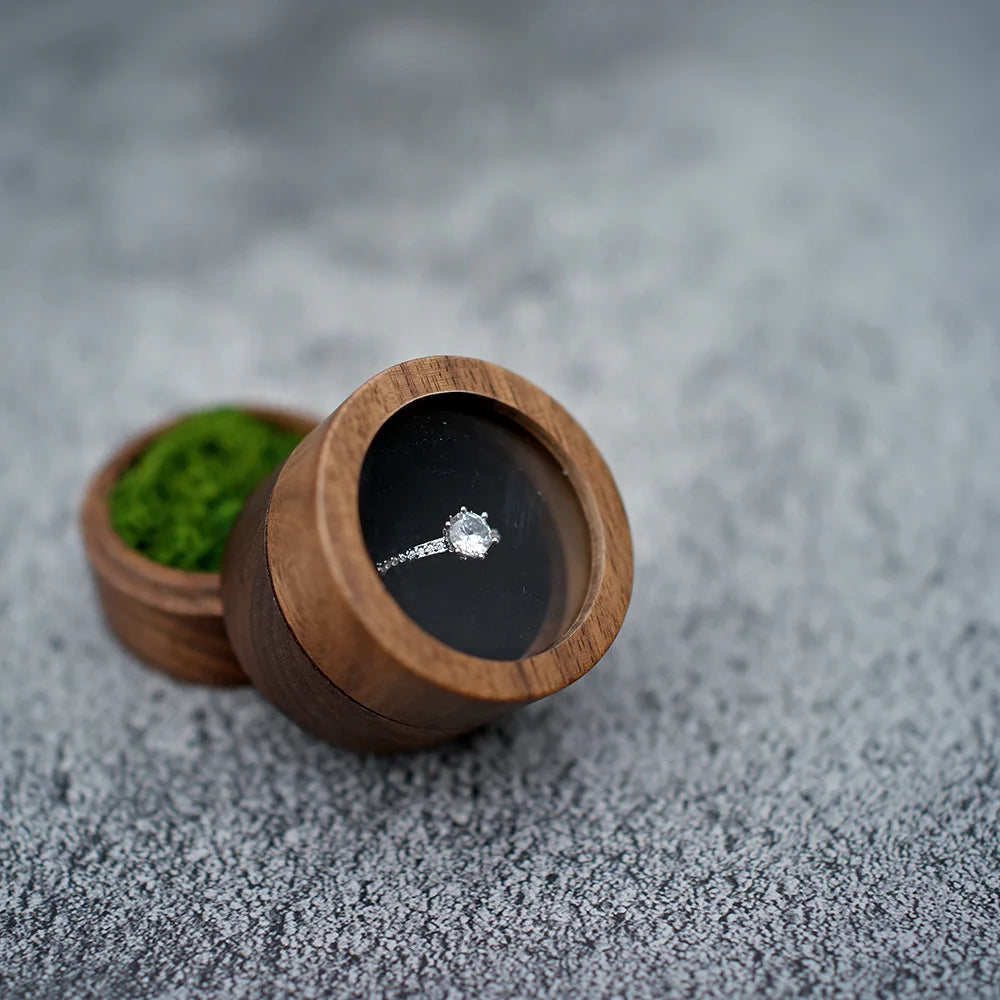 Boîte à bague de fiançailles ronde en bois sombre, ouverte, avec une bague en argent et diamant posée à l'intérieur. De la mo