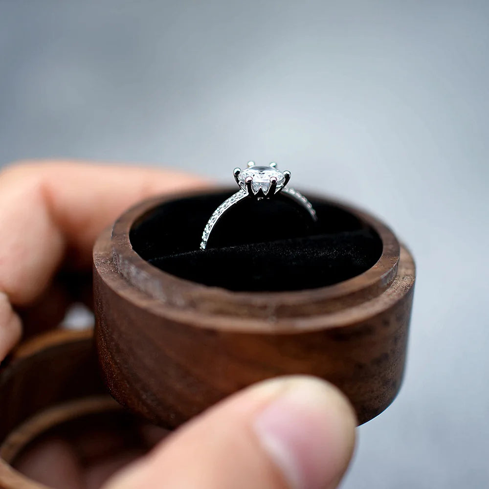 Une boîte à bague de fiançailles ronde en bois sombre, ouverte, tenant une bague en argent avec un diamant sur son coussin no