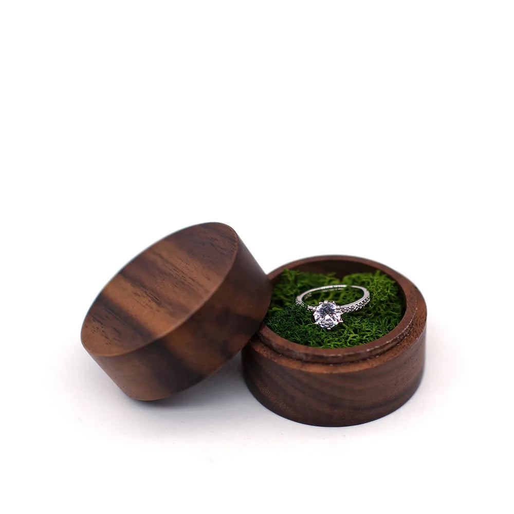 Boîte à bague de fiançailles ronde en bois sombre, ouverte. Une bague en argent avec pierre est posée sur de la mousse verte.