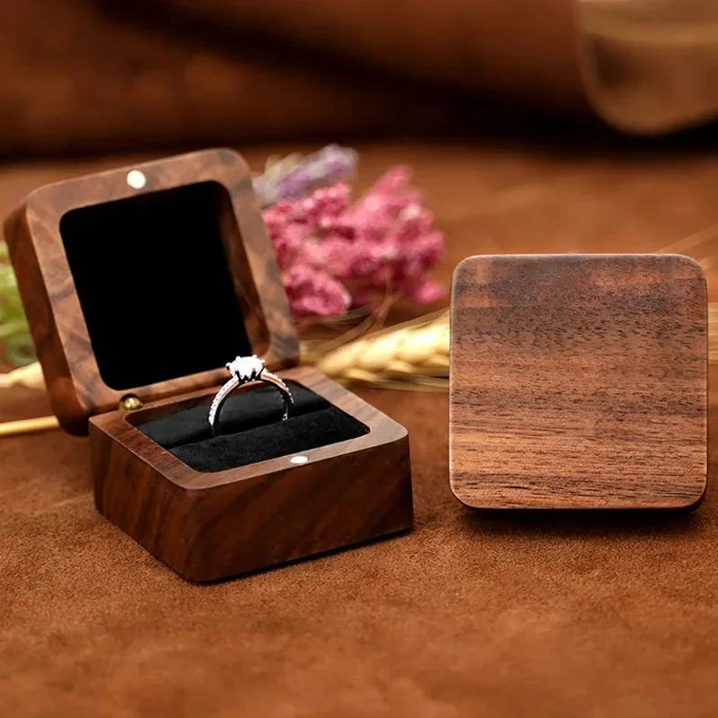 Boîte à bagues en bois brun foncé à l'aspect grainé, ouverte avec un anneau serti sur fond noir. Un couvercle est à côté.