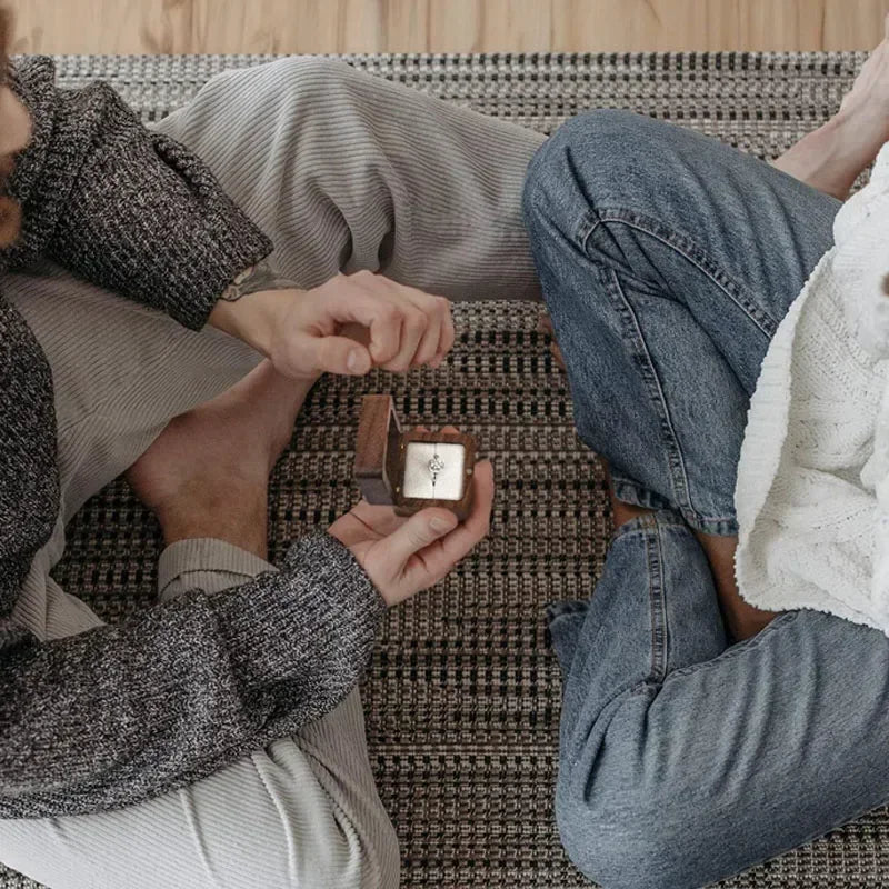 Boîte à bagues en bois de 5,5 cm avec couvercle ouvert, posée dans les mains d'une personne assise au sol.