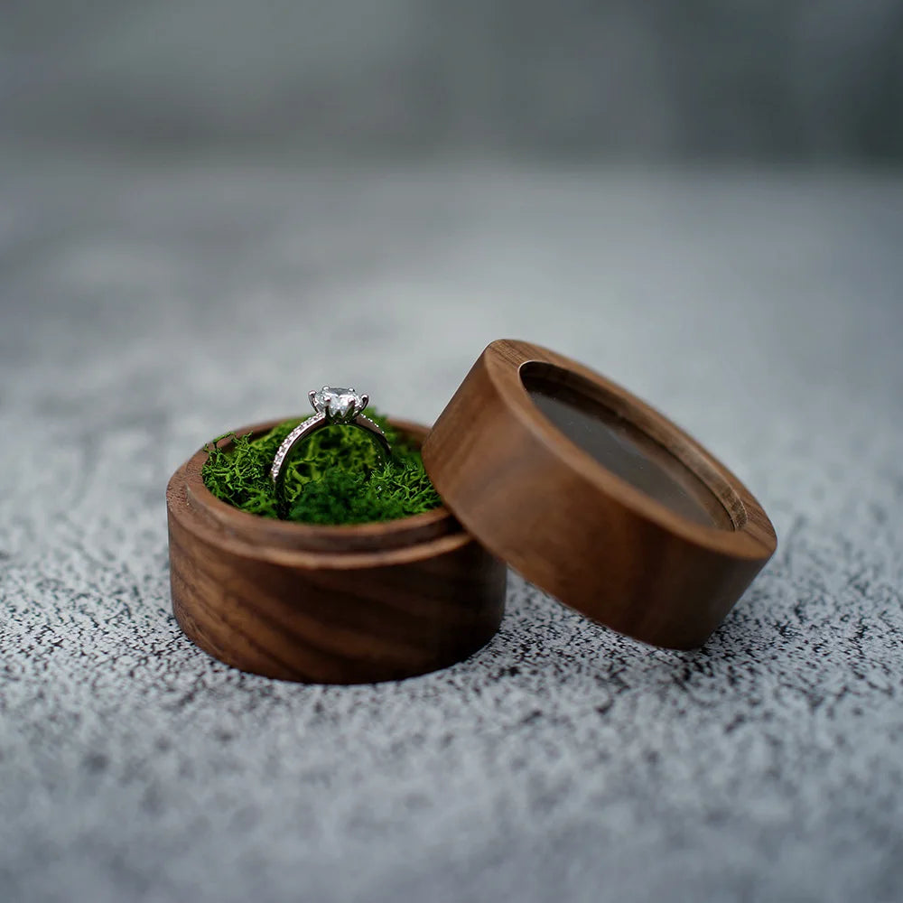 Boîte à bague de fiançailles ronde en bois sombre, ouverte. Un coussin de mousse verte maintient une bague en argent ornée.