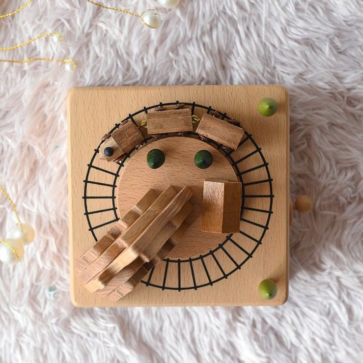 Boîte à musique carrée en hêtre, avec un train en bois gravé tournant sur des rails noirs et des accessoires.