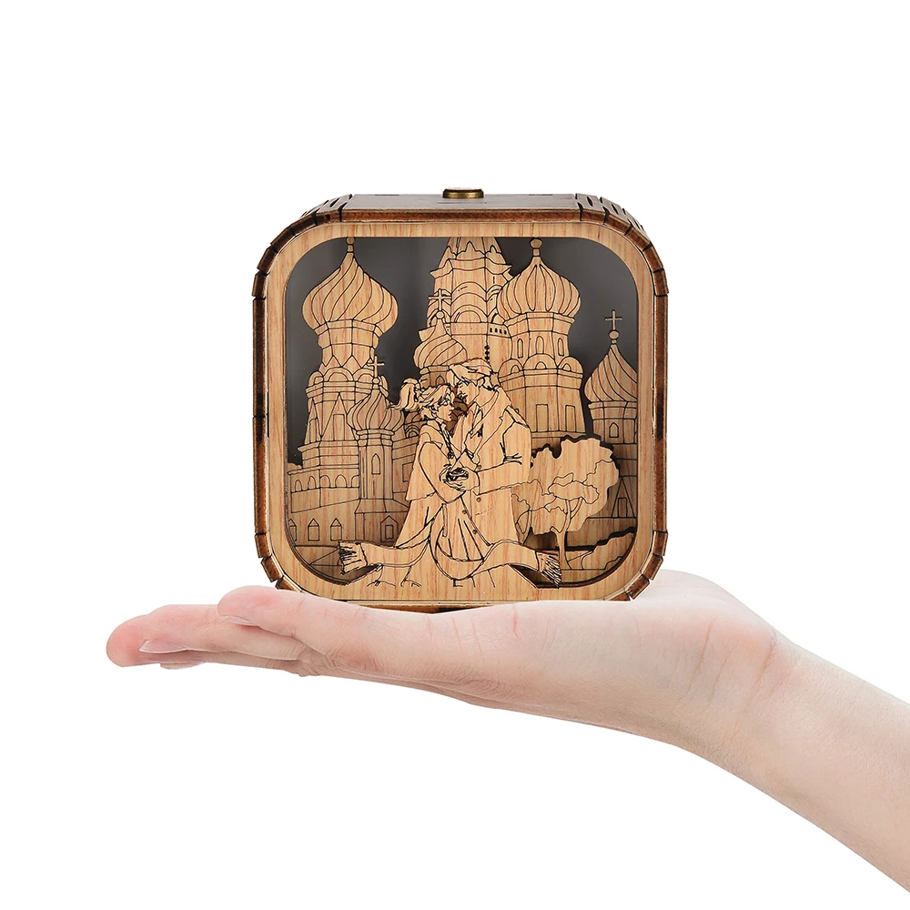 Boîte à musique veilleuse en bois, sur une main. Décoration gravée d'un couple devant une architecture. 10x10x8 cm.