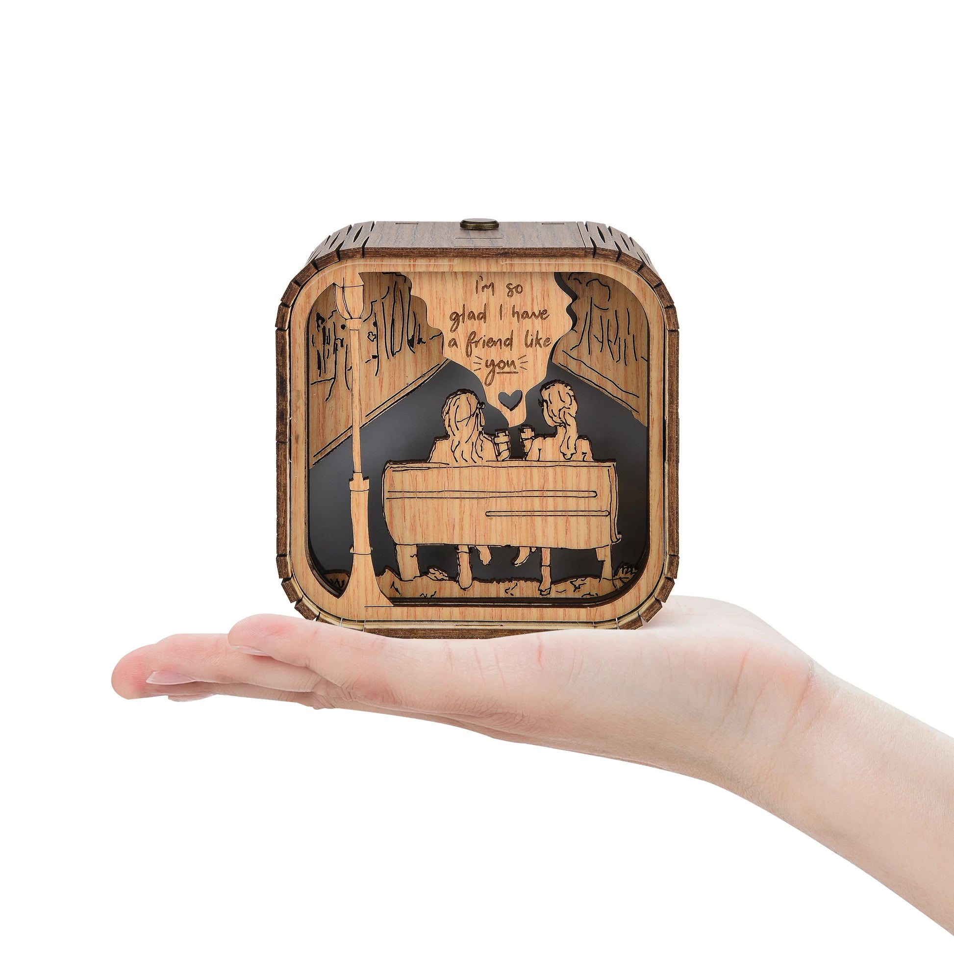 Boîte à musique en bois de 10x10x8cm sur une main ouverte, motif gravé d'amis et d'une lampe de rue.