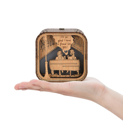 Boîte à musique en bois de 10x10x8cm sur une main ouverte, motif gravé d'amis et d'une lampe de rue.