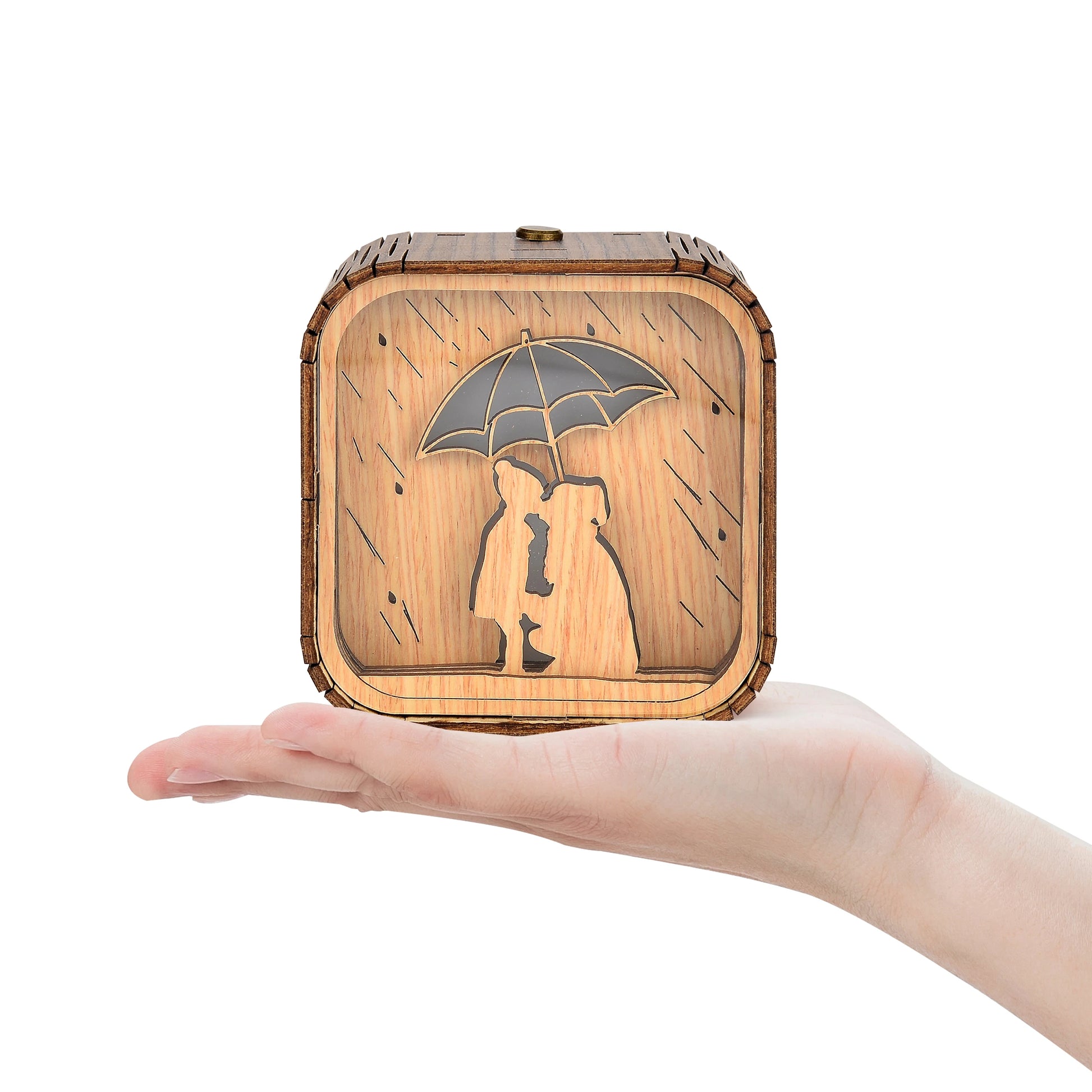 Boîte à musique veilleuse en bois, gravée d'un couple sous un parapluie avec des gouttes de pluie. Vue de face sur la paume d