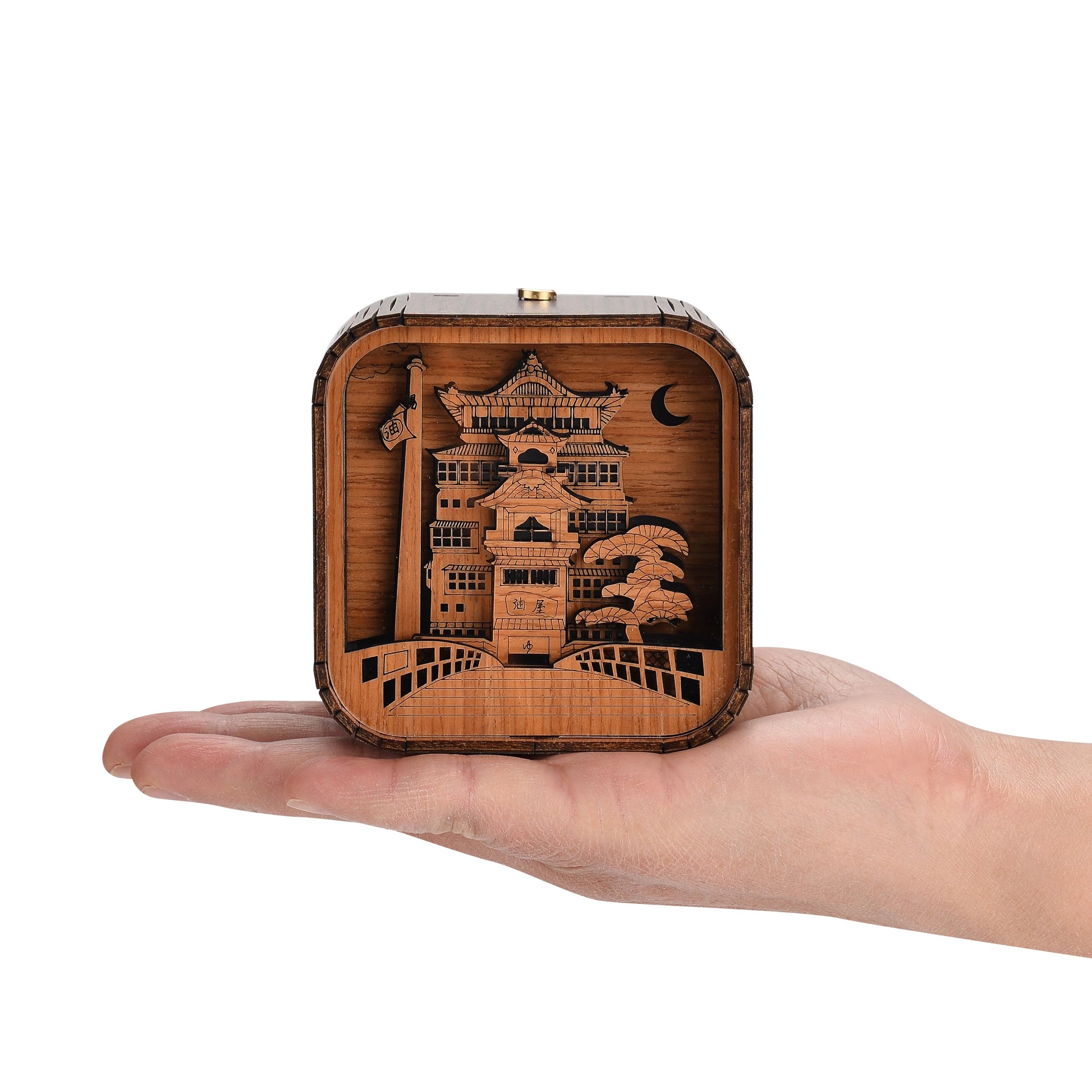 Boîte à musique veilleuse en bois, en forme de cube, montrant un bâtiment et des arbres découpés, tenue dans une main.