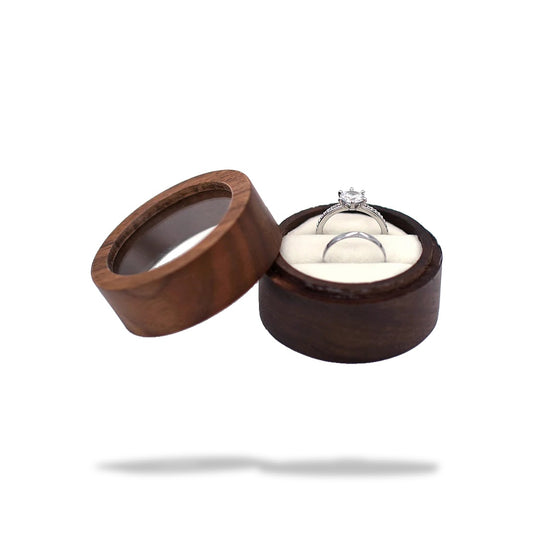 Boîte à bague de fiançailles ronde en bois sombre, ouverte. Elle contient deux bagues en argent sur un coussinet blanc.