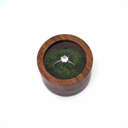 Boîte à bague de fiançailles ronde en bois avec couvercle transparent, contenant une bague argentée sur un lit de mousse vert