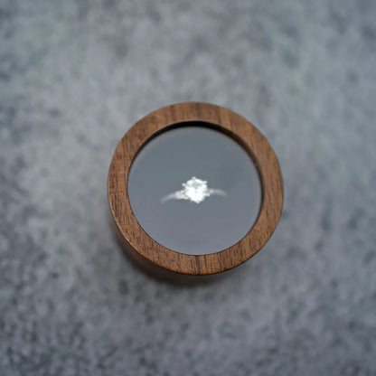 Boîte à bague de fiançailles ronde en bois sombre, avec couvercle transparent révélant une bague en argent.