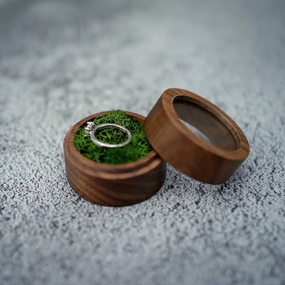 Boîte à bague de fiançailles ronde en bois avec couvercle séparé, intérieur en mousse verte tenant une bague en argent.