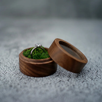 Boîte à bague de fiançailles ronde en bois sombre, ouverte. Un coussin de mousse verte maintient une bague en argent ornée.